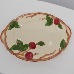Puede incluir: Plato ovalado para servir con un centro color crema y un borde marrón. El borde está decorado con ramas marrones, hojas verdes y fruta roja. El plato tiene un borde festoneado.