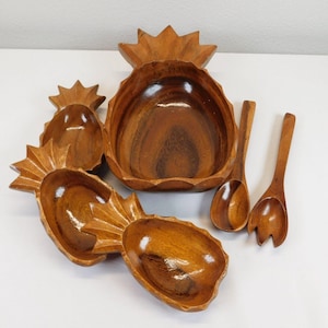 Puede incluir: Un juego de cinco cuencos de madera con forma de piña y dos utensilios para servir. Los cuencos están tallados en forma de piña, con el cuenco más grande en el centro y cuatro cuencos más pequeños que lo rodean. Los utensilios son una cuchara de madera y un tenedor de madera.