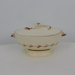 May include: Cream-colored, lidded serving dish with a pedestal base. The dish features brown and orange leaf-like designs and a gold rim. The lid has a small, decorative knob. Handles are on either side of the dish.