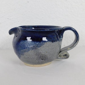 May include: A small, blue and gray ceramic creamer with a unique handle. The creamer has a speckled glaze and a slightly uneven shape.