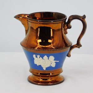 Op de afbeelding: Een koperen kan met een blauw en wit ontwerp. De kan heeft een rond lichaam met een gegolfde lip en een handvat. Het ontwerp heeft een witte cameo van een bloemmotief op een blauwe achtergrond.