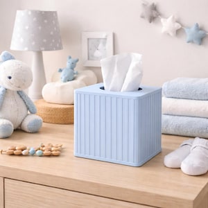 May include: A light blue, square tissue box with vertical ribbed detailing. White tissues are pulled from the top opening. The box sits on a wooden surface, alongside a plush toy, lamp, and baby items.