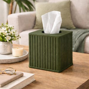May include: A square, olive green tissue box with vertical ribbed detailing. White tissues are pulled from the top opening. The box sits on a wooden table, with a candle, flowers, and books in the background. A decorative item for the home.