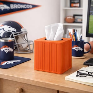 May include: An orange tissue box with vertical lines, filled with white tissues, sits on a wooden desk. A Denver Broncos theme is present with a mug, tumbler, helmet, and pennant in the background. The Broncos logo is visible on several items.