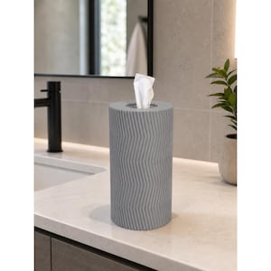 May include: A cylindrical gray tissue holder featuring a vertical chevron pattern embossed on its exterior surface. The holder has a circular opening at the top from which a single white tissue is protruding, providing a functional storage solution.