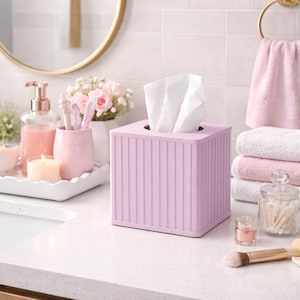 May include: A light pink square tissue box with vertical lines, holding white tissues. The box sits on a white countertop, with a gold-framed mirror, pink towels, and other bathroom accessories in the background.