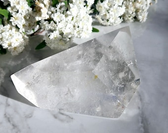 May include: A clear quartz crystal point with a double termination, also known as a double-ended crystal. The crystal is lying on a white marble surface.
