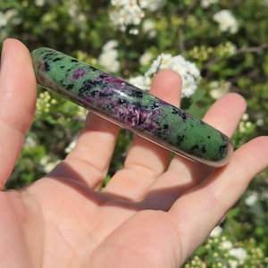 May include: A polished, elongated stone with a mix of green, black, and purple hues. The stone is held in a hand, with a blurred background of white flowers and green foliage. The stone's surface is smooth and reflective.