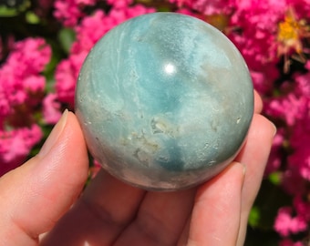 May include: A smooth, green and white agate sphere, polished to a high shine. The sphere is held in a hand, with a blurry background of pink flowers.