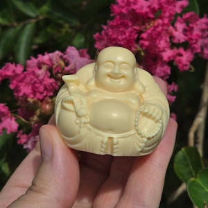May include: A small, white, ceramic figurine of a smiling Buddha holding a string of beads and a small cup. The figurine is sitting in a relaxed pose with its hands resting on its lap.