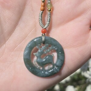 May include: A jade pendant necklace featuring a circular, dark green pendant with a carved deer silhouette. The pendant hangs from a gray cord with orange and gold-colored beads. The necklace is displayed against a blurred background.