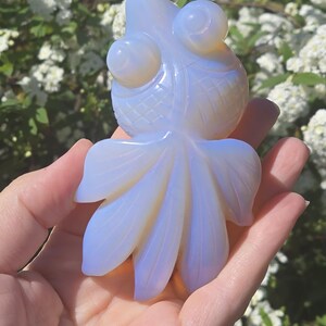 May include: A carved opal fish figurine with a white and blue iridescent color. The fish has large eyes and a detailed tail.