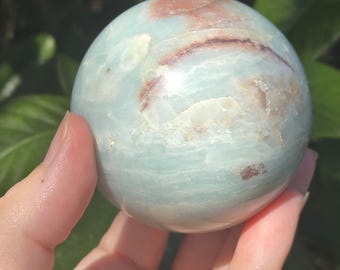 May include: A polished sphere of aquamarine and brown stone. The orb has a smooth surface with natural patterns and variations in color. The sphere is held in a hand, with a blurred green background of foliage.