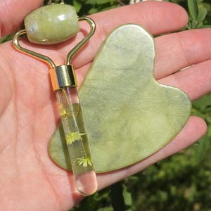 May include: A green jade facial roller and gua sha set. The roller has a gold handle and a clear tube filled with small white flowers. The gua sha is heart-shaped.