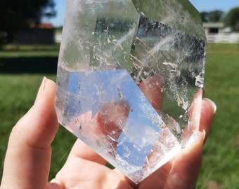 May include: A clear quartz crystal point with a double terminated shape. The crystal is held in a hand against a green grassy background.