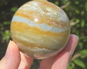 May include: A smooth, round, banded sphere of polished agate with shades of yellow, brown, and gray. The sphere is about the size of a golf ball.