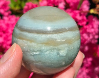 May include: A smooth, light blue and brown agate sphere with swirling patterns. The sphere is about the size of a golf ball and is being held in a hand.