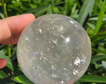 May include: A clear quartz crystal sphere with a cloudy, milky appearance. The sphere is smooth and polished, and it has a slight iridescent sheen.