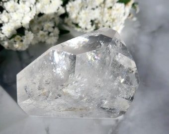 May include: A clear quartz crystal with a faceted shape. The crystal is sitting on a white marble surface.