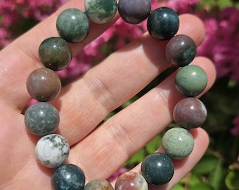 May include: A beaded bracelet featuring a variety of natural stone beads in shades of green, brown, and purple. The round beads are strung together, creating a colorful and earthy accessory. The bracelet is held in a hand against a backdrop of pink flowers.