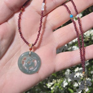 May include: A delicate necklace featuring a circular jade pendant with a carved design. The pendant hangs from a strand of small, dark red beads, accented with gold-colored findings and small, colorful beads. The necklace is displayed against a natural, outdoor background.