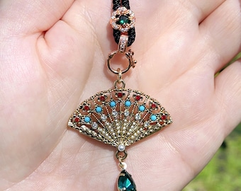 May include: A gold fan-shaped pendant necklace with red, blue, and white gemstones. The pendant is attached to a black cord and has a green teardrop-shaped gemstone hanging from the bottom.