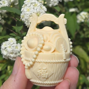 May include: Ivory-colored carved basket figurine with a handle, filled with various objects, including a coin and beads. The basket has a diamond-shaped emblem with a Chinese character. The figurine is held in a hand against a backdrop of white flowers.