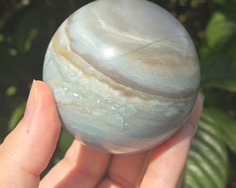 May include: A smooth, polished sphere of blue and white banded agate. The agate has a swirling pattern of light and dark blue bands with white accents.