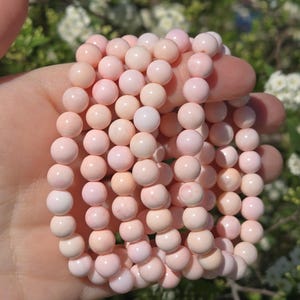 May include: A collection of light pink beaded bracelets. The beads are round and have a smooth, polished appearance. The bracelets are held in a hand, with a blurred background of green foliage and white flowers.