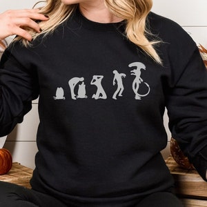 May include: Black sweatshirt with a gray graphic of the evolution of the alien from the movie Alien. The graphic shows a series of figures, starting with a primitive creature and ending with the iconic alien.