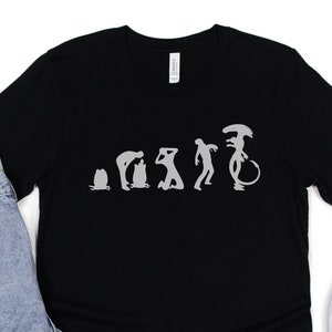 May include: Black t-shirt with a white graphic of the evolution of man ending with an alien. The shirt is displayed on a white background with a pair of white sneakers and blue jeans.