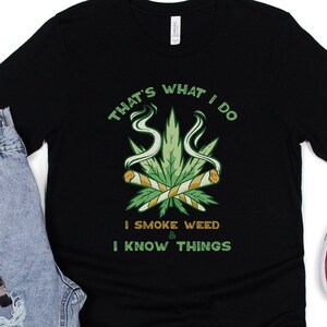 May include: Black t-shirt with the text "THAT'S WHAT I DO I SMOKE WEED & I KNOW THINGS" in green and white. The graphic features a cannabis leaf with two rolled joints. Light blue jeans, a watch, and cream-colored sneakers are also in the image.