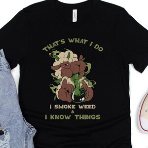 May include: Black t-shirt with a cartoon sheep smoking a bong, surrounded by green smoke. Text reads "THAT'S WHAT I DO I SMOKE WEED & I KNOW THINGS." The shirt is paired with light blue jeans, a watch, and beige sneakers.