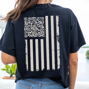 May include: A black t-shirt featuring a distressed white American flag design and a QR code on the back. The shirt is being worn by a person with long brown hair.