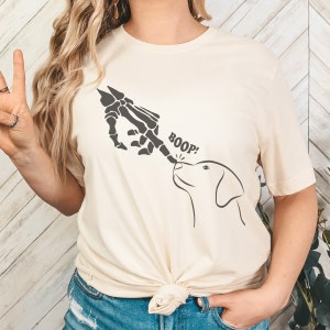 May include: A cream-coloured t-shirt with a black and white graphic of a skeleton hand booping a dog's nose. The text "BOOP!" is printed on the shirt.