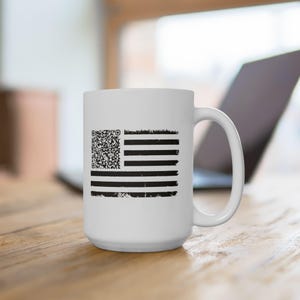 May include: White ceramic mug featuring a distressed black and white American flag design. The flag's stars are replaced with a QR code. The mug has a large handle and is set on a wooden surface.