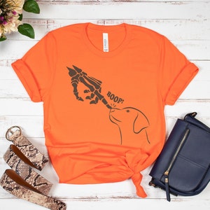 May include: Orange t-shirt featuring a skeleton hand booping a dog's nose. The text "BOOP!" is printed on the shirt.