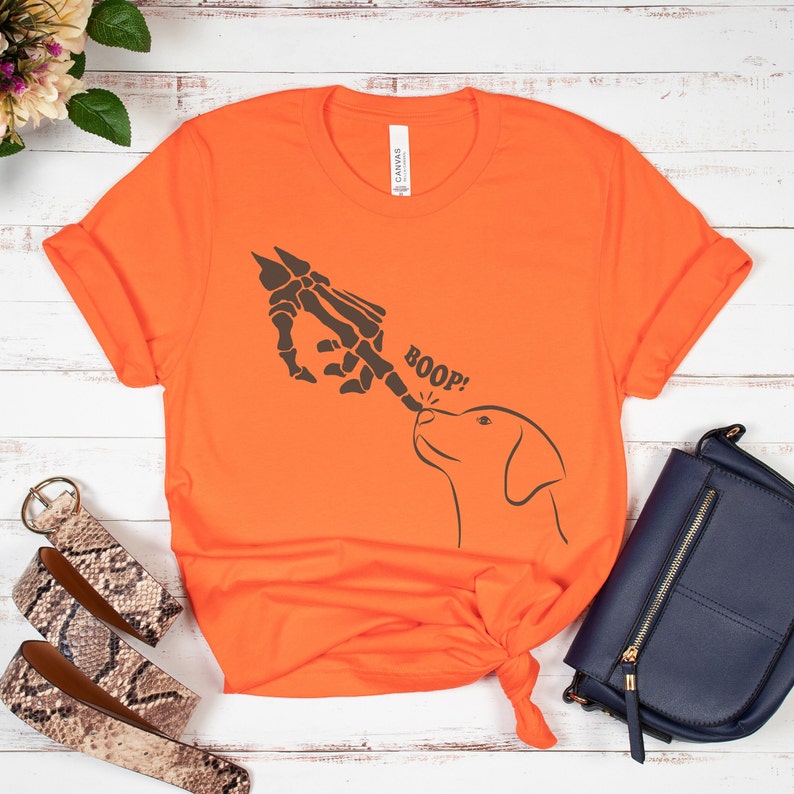May include: Orange t-shirt featuring a skeleton hand booping a dog's nose. The text "BOOP!" is printed on the shirt.