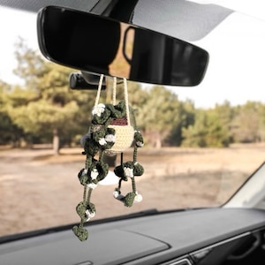 May include: A crocheted hanging plant car charm. The charm features a beige pot with a brown interior, and green and white crocheted leaves. The charm is suspended from a rearview mirror inside a car, with a blurred outdoor background.