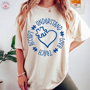 May include: A cream-colored short-sleeve t-shirt featuring a blue circular graphic. The design includes the words UNDERSTAND, LOVE, TEACH, and ACCEPT arranged in a circle around a heart shape with a puzzle piece cutout and small puzzle pieces.