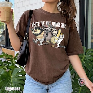 May include: A brown short-sleeve t-shirt featuring the text "WHERE THE WILD THINGS ARE" printed in white above a graphic illustration of four monsters from the story. The shirt has a crew neckline and a relaxed fit, shown in a solid brown colour.