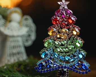 May include: A rainbow-colored Christmas tree ornament made of rhinestones. The tree is decorated with a star on top and is about 6 inches tall.