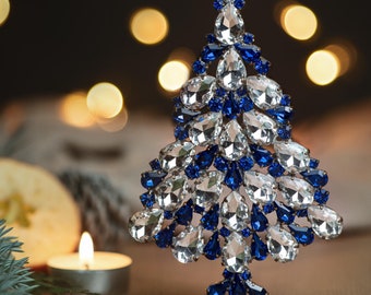 May include: A blue and clear crystal Christmas tree ornament with a star on top. The ornament is on a gold stand and is surrounded by a blurred background of lights.
