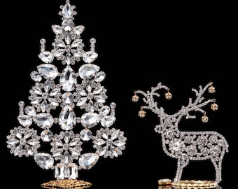 May include: A sparkling Christmas tree and reindeer figurine set. The tree is adorned with clear, faceted crystals and sits on a gold-colored base. The reindeer is also embellished with crystals and has small, gold-colored orbs on its antlers.
