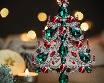 Puede incluir: Un adorno de árbol de Navidad brillante de color verde y rojo con joyas. El árbol está hecho de metal y tiene una base plateada.