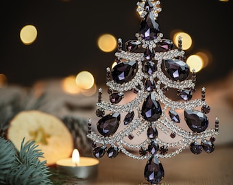 May include: A sparkling purple and silver Christmas tree ornament with a jeweled design. The ornament is on a gold stand and is surrounded by a blurred background of lights.