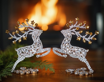 May include: Two silver reindeer figurines with gold accents and rhinestones, standing on a wooden surface in front of a fireplace with a fire burning.