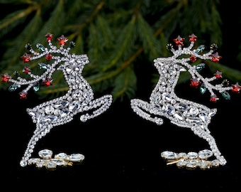May include: Two silver reindeer ornaments decorated with rhinestones and red and green crystals. The reindeer are in a leaping pose.