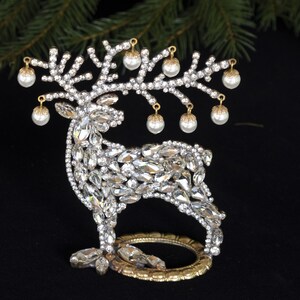 May include: A decorative reindeer figurine made of rhinestones and gold metal. The reindeer has a gold metal base and is adorned with white pearl-like ornaments.