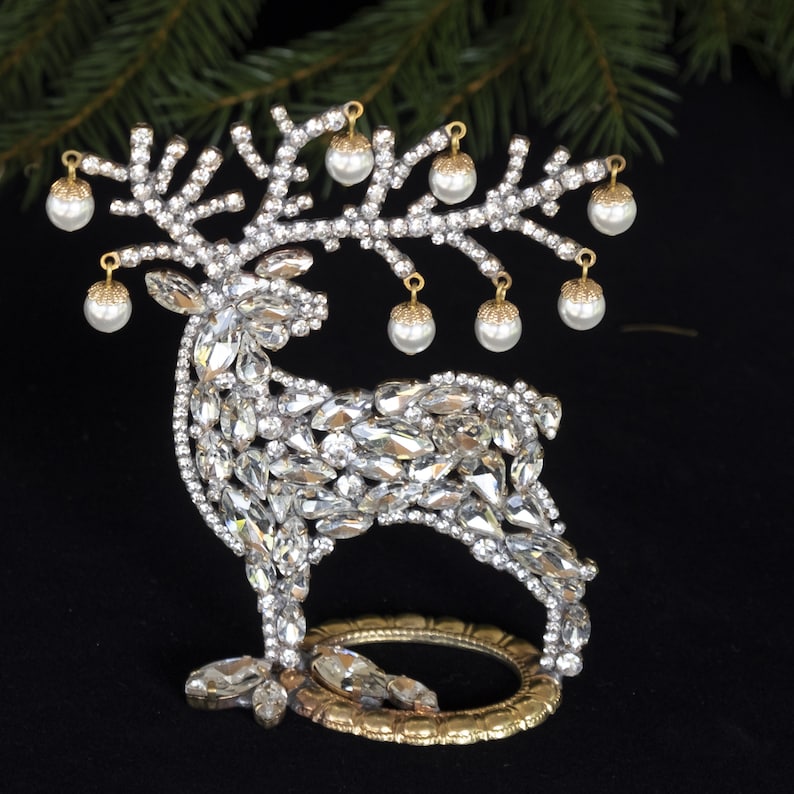 May include: A decorative reindeer figurine made of rhinestones and gold metal. The reindeer has a gold metal base and is adorned with white pearl-like ornaments.